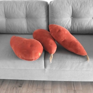 以下が含まれることがあります： グレーのソファに、オレンジがかった赤色のサツマイモ型のぬいぐるみクッションが3つ置かれています。クッションは柔らかくビロードのような質感で、端には小さなベージュのタッセルが付いています。ソファはボタン留めデザインです。