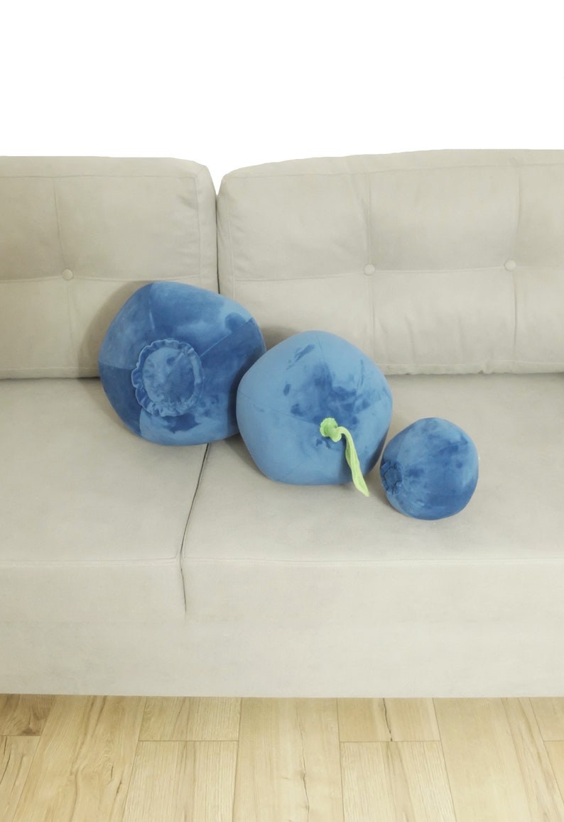 May include: Three plush blueberry pillows in different sizes on a light gray couch. The pillows are blue with green stems.