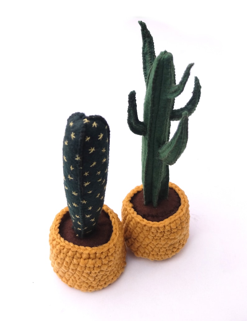 May include: Two potted felt cactus plants. The cactus on the left is a small, green cactus with yellow stars. The cactus on the right is a tall, green cactus with multiple arms. Both cacti are in yellow crocheted pots.
