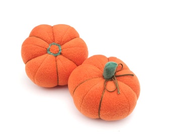 May include: Two orange plush pumpkins with green stems. The pumpkin on the left has a simple design, while the pumpkin on the right has a brown ribbon tied around the stem.