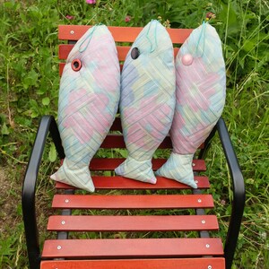 Peut inclure: Trois coussins en forme de poisson de couleur pastel avec des yeux en boutons sont pos&eacute;s sur un banc en bois. Les coussins sont faits d'un tissu doux et &agrave; motifs et ont un aspect artisanal et fantaisiste.
