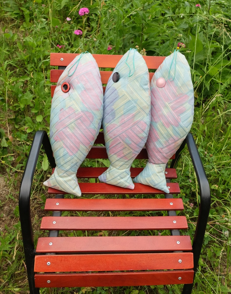 Peut inclure: Trois coussins en forme de poisson de couleur pastel avec des yeux en boutons sont pos&eacute;s sur un banc en bois. Les coussins sont faits d'un tissu doux et &agrave; motifs et ont un aspect artisanal et fantaisiste.