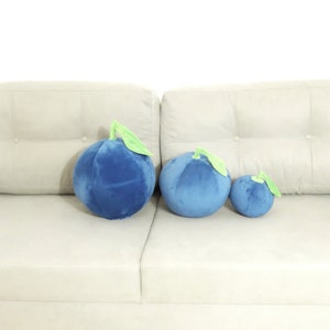 May include: Three plush blueberry pillows of different sizes on a white couch. The largest pillow is on the left, the medium-sized pillow is in the middle, and the smallest pillow is on the right.