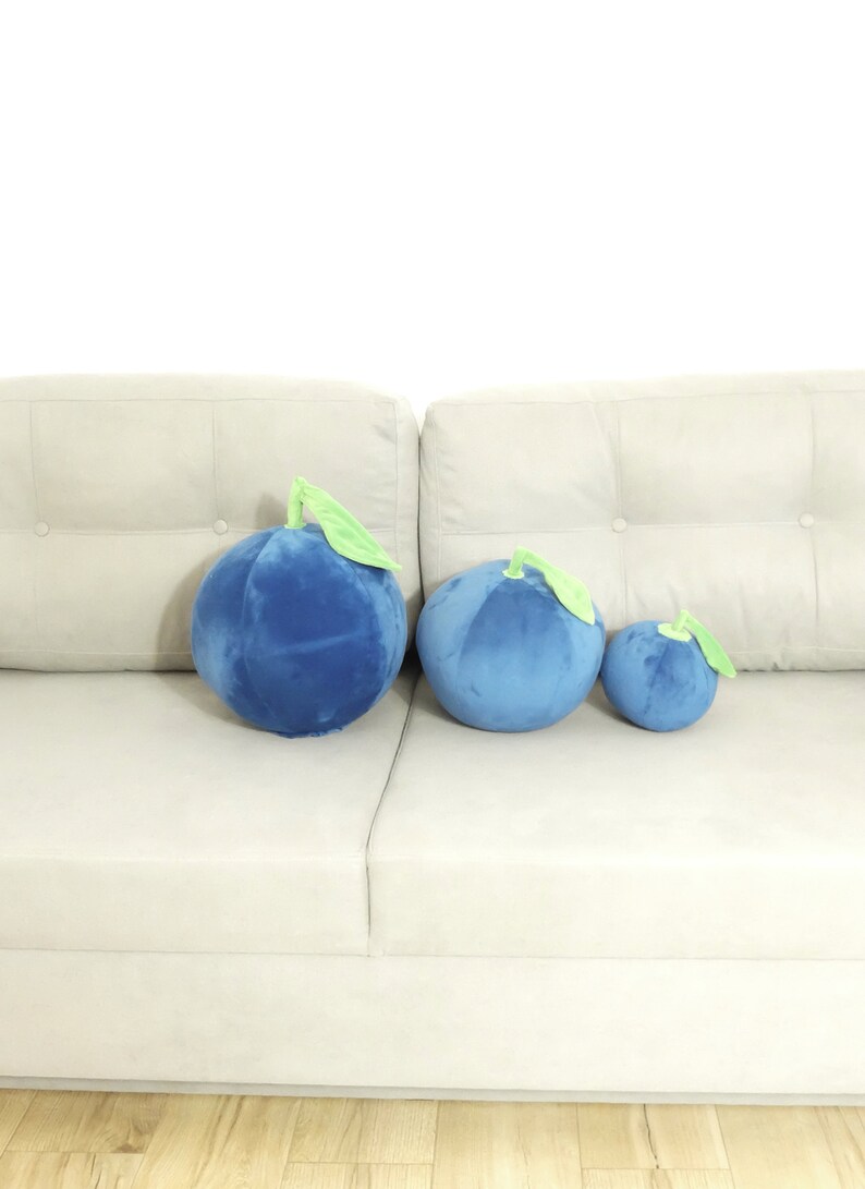 May include: Three plush blueberry pillows of different sizes on a white couch. The largest pillow is on the left, the medium-sized pillow is in the middle, and the smallest pillow is on the right.