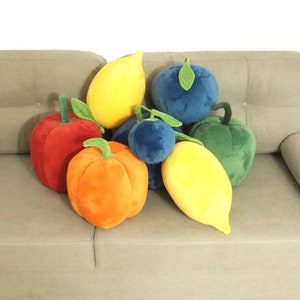 May include: A group of plush fruit pillows in various colors, including red, orange, yellow, green, and blue. The pillows are arranged on a light gray couch.
