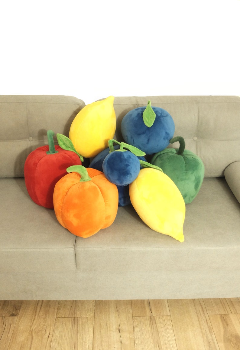 May include: A group of plush fruit pillows in various colors, including red, orange, yellow, green, and blue. The pillows are arranged on a light gray couch.