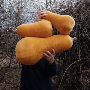 Könnte beinhalten: Eine Person hält drei große, plüschige, orangefarbene Butternusskürbis-Kissen. Die Kissen sind aus einem weichen, samtigen Stoff.