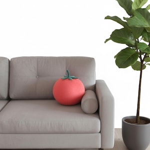 Può includere: Un cuscino rotondo a forma di pomodoro, color corallo, con uno stelo verde, poggia su un divano grigio chiaro. Il divano ha un cuscino cilindrico abbinato ed è accanto a una pianta in vaso.