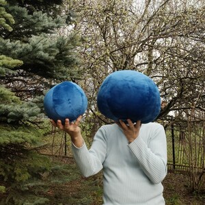 May include: Two blue plush balls, one larger than the other, are held in front of a person's face. The person is wearing a light blue sweater.