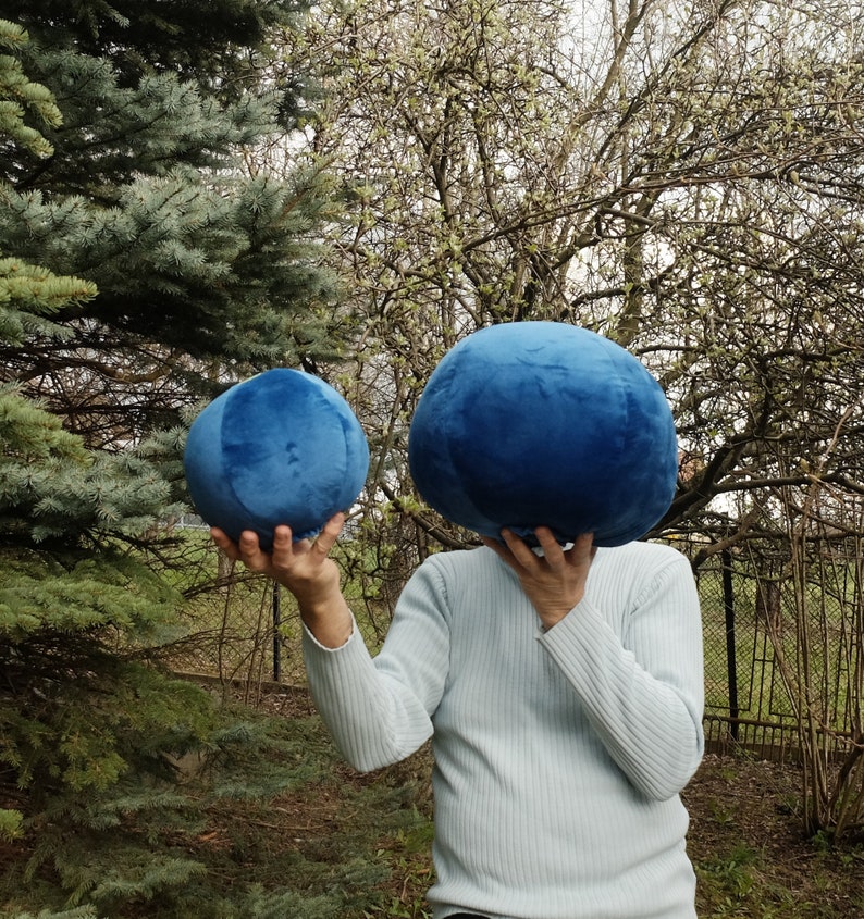 May include: Two blue plush balls, one larger than the other, are held in front of a person's face. The person is wearing a light blue sweater.