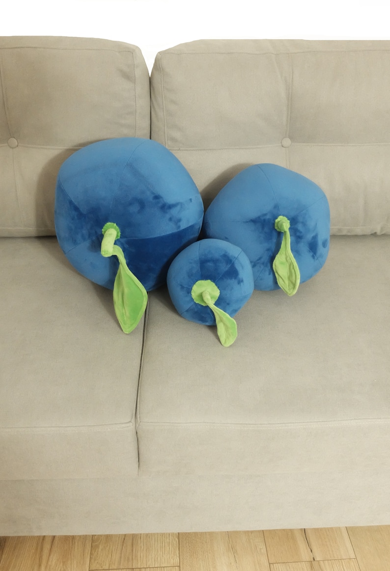 May include: Three plush blueberry pillows in different sizes, with green stems and leaves, on a light gray couch.