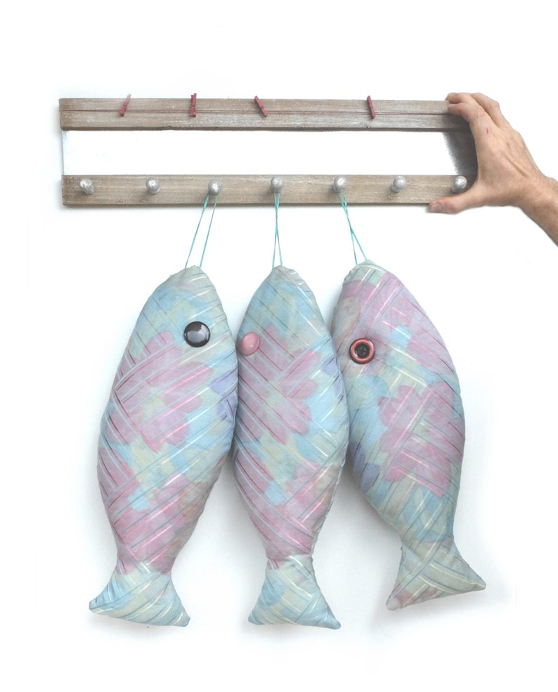 Peut inclure: Trois ornements en forme de poisson de couleur bleu pastel et rose suspendus &agrave; un support en bois. Chaque poisson a un &oelig;il en bouton et est fait d'un tissu tiss&eacute;.