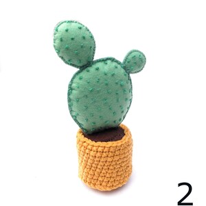 May include: A green felt cactus with a brown felt base, sitting in a yellow crocheted pot. The cactus has two round pads and is stitched with green thread.
