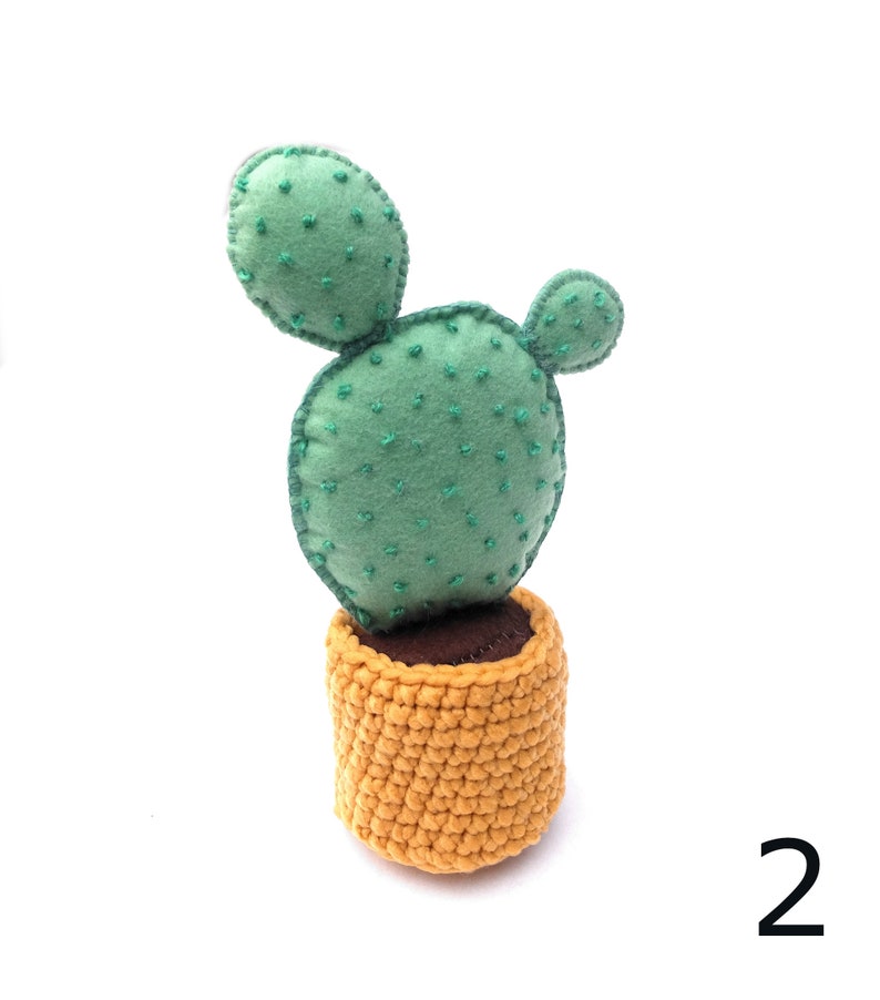 May include: A green felt cactus with a brown felt base, sitting in a yellow crocheted pot. The cactus has two round pads and is stitched with green thread.