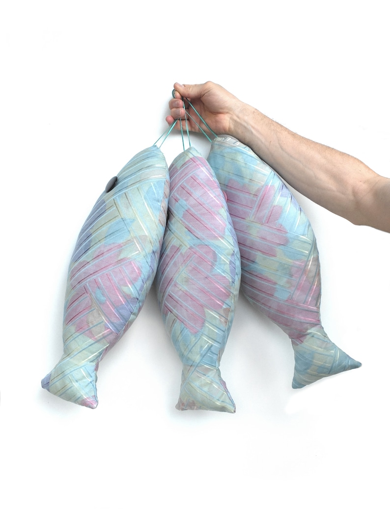 Peut inclure: Trois coussins en forme de poisson &agrave; motifs bleus et roses sont suspendus &agrave; une ficelle tenue par une main. Les coussins sont faits d'un tissu doux et textur&eacute;.