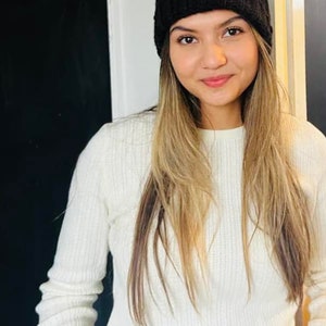 Op de afbeelding: Een vrouw met lang blond haar die een witte trui en een zwarte gebreide beanie draagt.