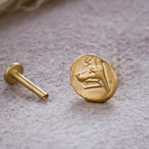 May include: A gold-colored stud earring with a flat, round face featuring a detailed relief of a face. The earring post is a slender, cylindrical shape. The background is a soft, textured gray surface with a blurred, beige accent.