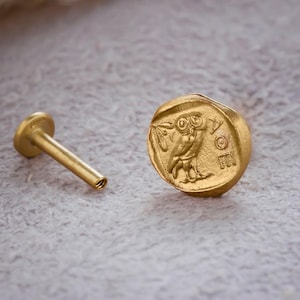 Pode incluir: Um piercing labial dourado. Apresenta um design plano e redondo com um emblema de coruja. Possui um poste reto e uma parte traseira plana. A joia é colocada sobre um fundo neutro.