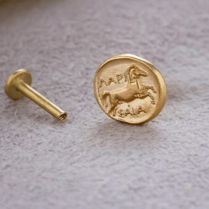 May include: A gold-colored stud earring with a flat, circular design featuring a horse and Greek text. The earring post is visible next to the circular design. The background is a light gray color.