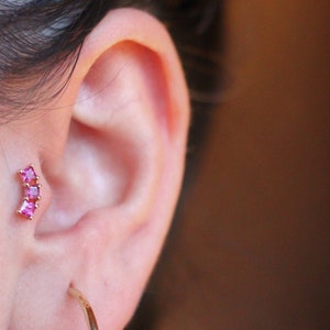 Pode incluir: Um close-up de uma orelha com um pequeno brinco de ouro com pedras preciosas rosa. O brinco está na piercing do hélix. A pessoa está usando um brinco de argola de ouro no lóbulo inferior.