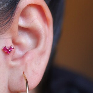 May include: A close-up of a person's ear with a small heart-shaped earring featuring pink gemstones. The earring is in the helix piercing. The person is also wearing a gold hoop earring in their lower lobe.