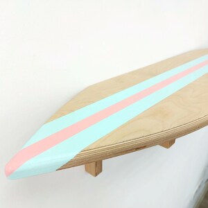 May include: A wooden surfboard shelf painted with light blue and pink stripes. The shelf is mounted to a wall and has a rounded edge.