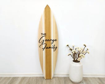 Op de afbeelding: Een hoge, puntige houten surfplank wanddecoratie met verticale licht- en middenbruine houtnerfpanelen. De tekst "THE George Family" staat in zwarte, gestileerde letters in het midden van het bord. Een witte vaas met gedroogde bloemen staat ernaast.
