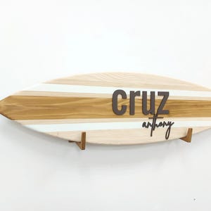 Puede incluir: Arte de pared con forma de tabla de surf de madera clara, acentuada por rayas en azul claro y marrón. El nombre "Cruz" se muestra en letras marrones oscuras, con "anthony" debajo en una fuente cursiva.