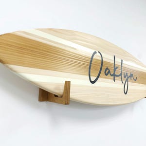 May include: A wooden surfboard with a natural wood grain finish and white stripes. The surfboard is mounted on a wall with two wooden brackets. The name "Oaklyn" is written in black cursive on the surfboard.