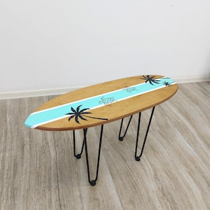 May include: A surfboard-shaped coffee table with a light brown wood grain finish. A central horizontal turquoise stripe runs across the surface, decorated with black silhouettes of palm trees and two sea turtles. It has four black metal hairpin legs.