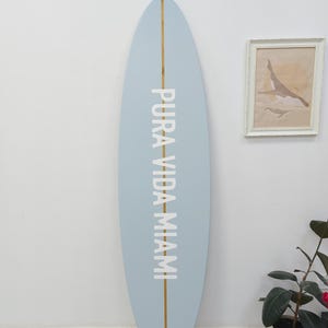 K&ouml;nnte beinhalten: Ein hellblaues Surfbrett mit einem vertikalen Mittelstreifen aus Holz und dem wei&szlig;en Schriftzug PURA VIDA MIAMI, der vertikal in der Mitte aufgedruckt ist. Das Surfbrett hat eine abgerundete Nase und ein abgerundetes Heck mit matter Oberfl&auml;che.