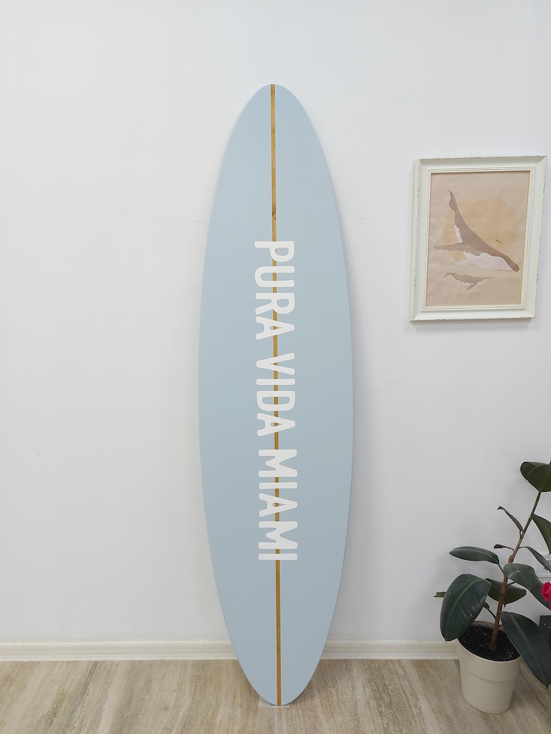 K&ouml;nnte beinhalten: Ein hellblaues Surfbrett mit einem vertikalen Mittelstreifen aus Holz und dem wei&szlig;en Schriftzug PURA VIDA MIAMI, der vertikal in der Mitte aufgedruckt ist. Das Surfbrett hat eine abgerundete Nase und ein abgerundetes Heck mit matter Oberfl&auml;che.