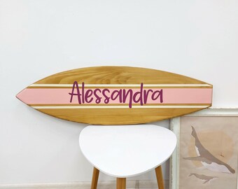 May include: A wooden surfboard-shaped wall sign featuring a natural wood grain finish. A horizontal light pink stripe runs across the center, bordered by thin white lines. The name Alessandra is printed in a dark purple script font.