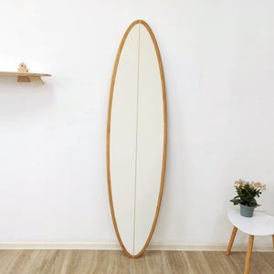Pode incluir: Um espelho alto de formato oval com uma moldura de madeira em tom claro. O espelho apresenta um painel central branco liso dividido por uma linha vertical fina, envolto numa borda de madeira arredondada que segue uma silhueta alongada.