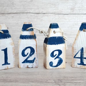 Peut inclure: Quatre décorations en bois en forme de bouée, peintes en blanc avec des rayures bleues et les chiffres 1, 2, 3 et 4. Chaque bouée a une corde attachée en haut. Le fond est un grain de bois clair.