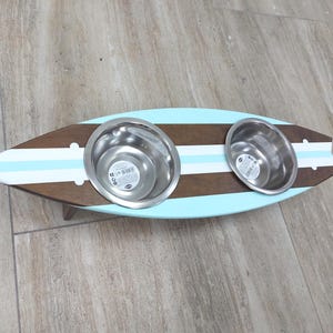 Può includere: Un supporto per ciotole per cani in legno a forma di tavola da surf. Presenta due ciotole in acciaio inossidabile inserite in un design a strisce marroni, azzurre e bianche. Il supporto è rialzato da piccole gambe di legno.