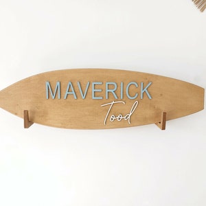 Könnte beinhalten: Ein surfboardförmiges Wand-Schild aus Holz mit dem Namen MAVERICK in hellblauen Blockbuchstaben und dem Namen Tood in weißer Schreibschrift. Das Schild ist auf zwei kleinen Holzhalterungen montiert und zeigt eine natürliche Holzmaserung.