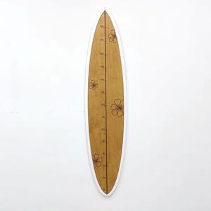 May include: A wooden surfboard-shaped growth chart with a white border. The chart has a light brown background with etched floral designs and height markings in inches. The surfboard is mounted on a white wall.