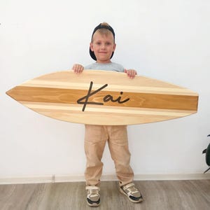 May include: A surfboard-shaped wooden balance board featuring a natural light and medium brown striped wood grain pattern. The centre of the board displays the name Kai in a black, cursive, handwritten-style font across the horizontal surface.