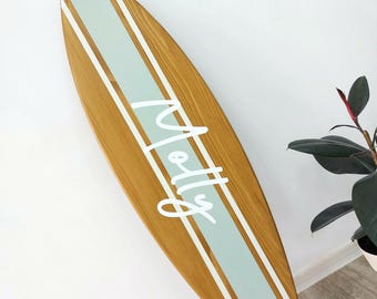 May include: A wooden surfboard featuring a natural brown wood grain finish with a wide, vertical light sage green stripe running down the centre, bordered by thin white lines. The name Molly is written in white script across the centre stripe.