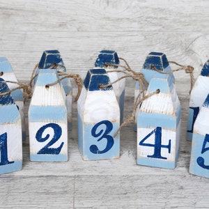 Op de afbeelding: Een slinger van houten boei-vormige ornamenten, beschilderd in wit, lichtblauw en marineblauw. Elke boei is genummerd van 1 tot en met 5 in marineblauw. De boeien zijn aan elkaar geregen met touw, wat een nautisch thema creëert.