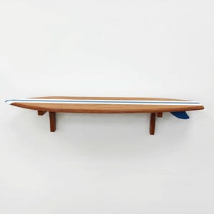 May include: A wooden surfboard shelf with a blue and white stripe design. The shelf is mounted to a white wall.