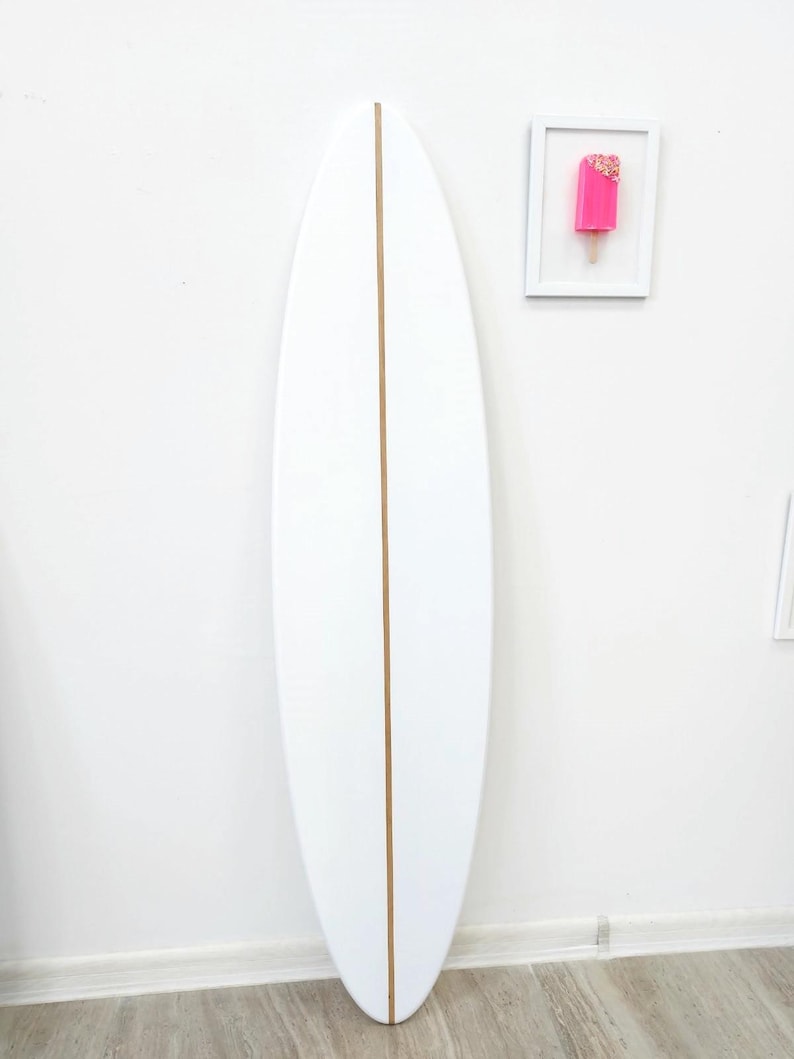 K&ouml;nnte beinhalten: Ein einfarbig wei&szlig;es Surfbrett mit einem d&uuml;nnen, vertikalen hellbraunen Holzstringer in der Mitte. Rechts daneben befindet sich ein kleines, wei&szlig; gerahmtes Kunstwerk, das ein leuchtend pinkes Eis am Stiel mit bunten Streuseln zeigt.