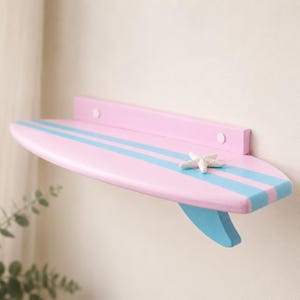 Puede incluir: Una estantería de pared rosa con forma de tabla de surf con dos rayas horizontales de color azul claro en la superficie. Una pequeña estrella de mar blanca está fijada en la parte superior, que también tiene una aleta azul claro debajo.