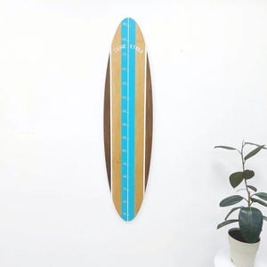 May include: Wooden growth chart in a surfboard shape. The chart features a light brown center with a blue stripe and white markings for height measurements in inches. The sides are a darker brown, and the text "GURE ETXEA" is printed at the top.