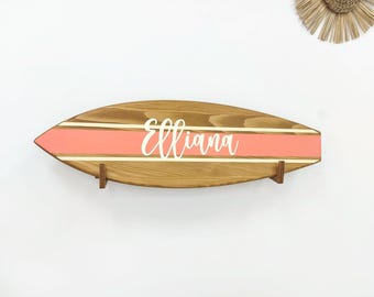 May include: A wooden surfboard-shaped wall sign featuring a horizontal coral-colored stripe flanked by thin white lines. The name Elliana is written in white cursive script across the center of the coral stripe. The sign is mounted on two small wooden brackets.