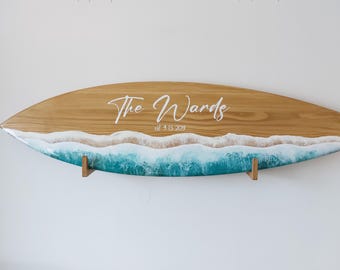 May include: A wooden surfboard with a blue and white ocean wave design. The text "The Wards est 9.13.2019" is written on the surfboard.