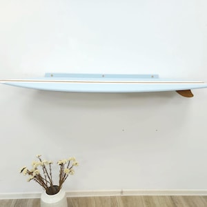 Könnte beinhalten: Hellblaues Surfbrett-Regal mit hellbrauner Finne und dünnem Goldstreifen. Das Regal ist an einer weißen Wand befestigt. Das Regal ist etwa 183 cm lang.