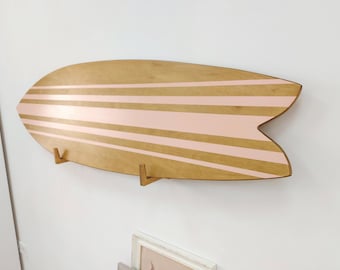 Puede incluir: Una estantería de pared con forma de tabla de surf de madera con un acabado de veta de madera marrón claro natural y rayas horizontales de color rosa pálido. La tabla tiene una punta afilada y una cola de golondrina, montada horizontalmente.