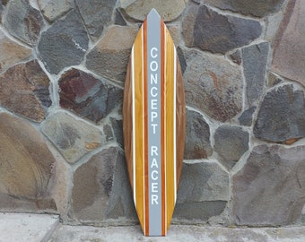 Op de afbeelding: Een houten surfboard met een bruin, geel en wit ontwerp. De tekst "CONCEPT RACER" is verticaal in het wit gedrukt op een grijze streep in het midden. Het surfboard wordt gepresenteerd tegen een stenen muur.